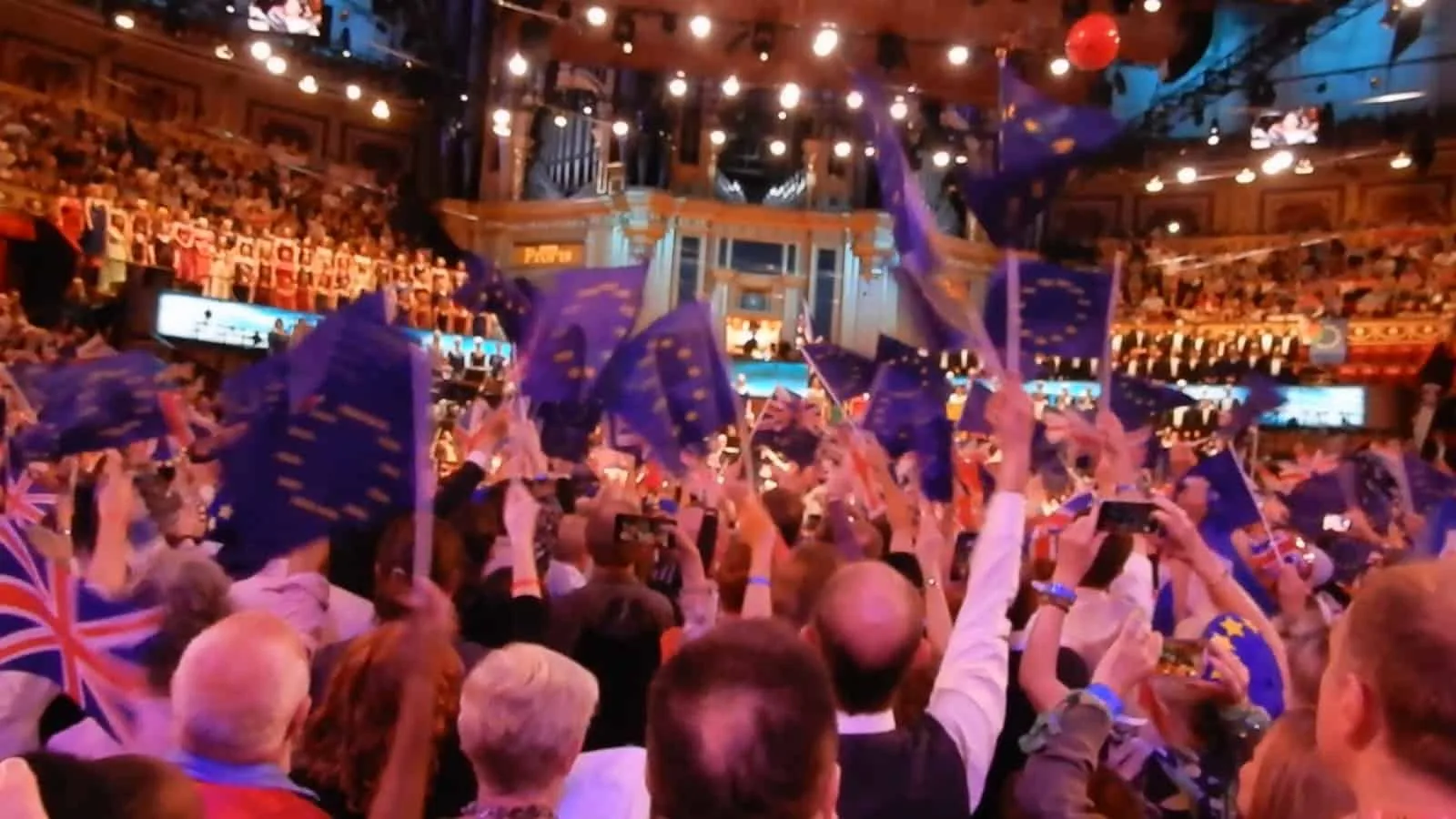 A large crowd of people is celebrating with Brexit flags and waving Union Jack flags in a grand, ornate indoor venue decorated with string lights.